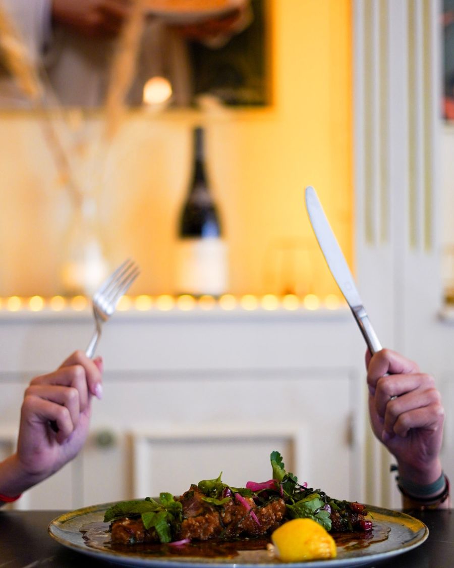 Deux mains tiennent une fourchette et un couteau au-dessus d'une assiette dressée, dans une salle de restaurant à la lumière chaude.